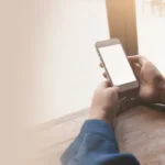 Person hält ein Smartphone in den Händen, sitzend am Tisch am Fenster.